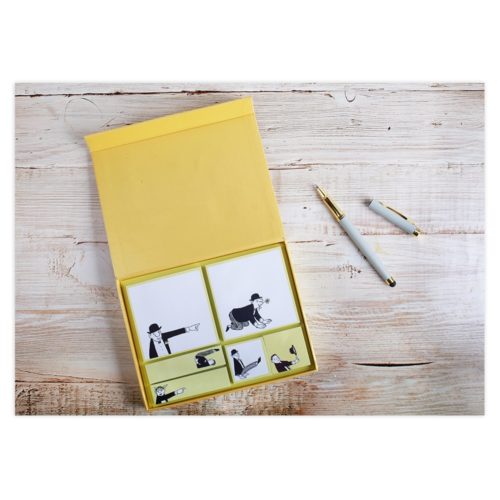 Offene gelbe Schachtel mit illustrierten Karten, neben einem Stift, auf einer Holzoberfläche.