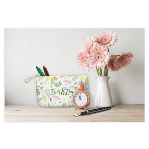 Rosa Gerbera in einer weißen Vase, ein Blumenbeutel mit Stiften, eine kleine Uhr und ein schwarzer Stift auf einer Holzoberfläche.