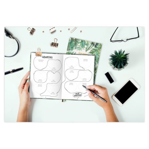 Hände schreiben in einen Planer auf einem Schreibtisch mit einem Telefon, einer Brille, einer Pflanze, Büroklammern und einem Stift.