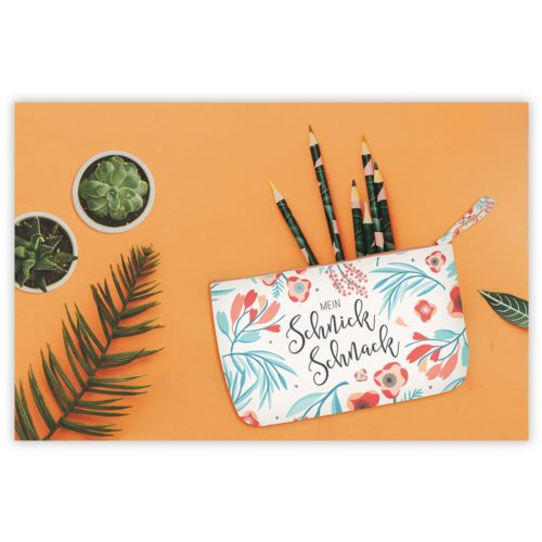 Federmäppchen mit Blumenmuster und Bleistiften auf orangefarbenem Untergrund, neben kleinen Sukkulenten und einem Palmblatt.