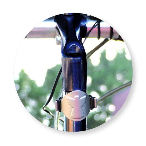 Nahaufnahme eines Fahrradlenkers mit einem daran befestigten kleinen engelförmigen Ornament, Bäume im unscharfen Hintergrund.