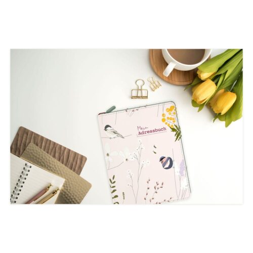 Ein geblümtes Adressbuch auf einem Schreibtisch mit Notizbüchern, Bleistiften, einer Tasse Kaffee und gelben Tulpen.