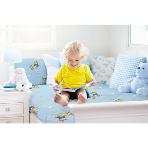 Ein Kind in einem gelben Hemd liest ein Buch auf einem blauen Bett mit Stern- und Tiermustern, neben einem Teddybären.