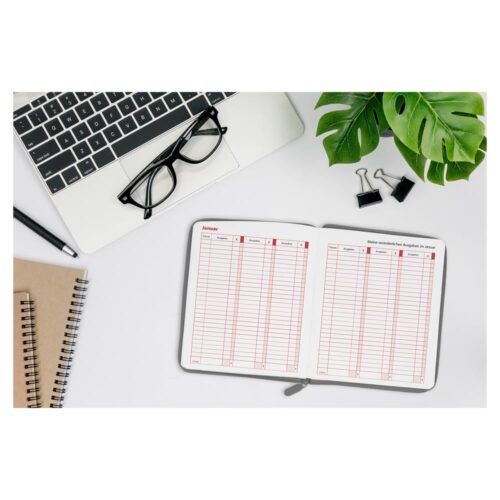 Offener Planer auf einem Schreibtisch mit Laptop, Brille, Notizbüchern, grünen Blättern und Heftklammern.