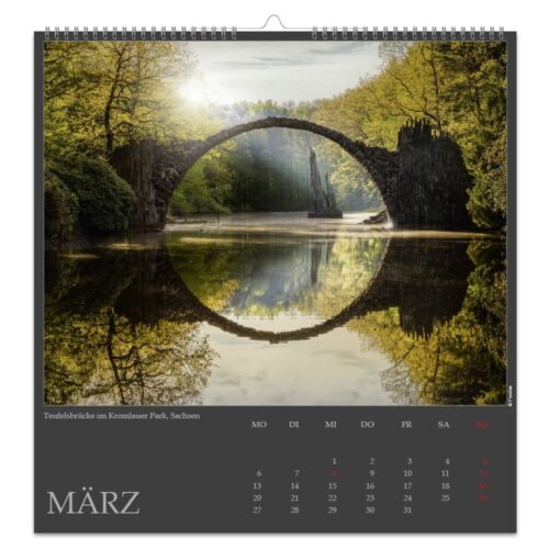 Malerische Aussicht auf die Rakotzbrücke, eine Steinbogenbrücke, die sich im Wasser spiegelt, umgeben von üppigen grünen Bäumen im März.
