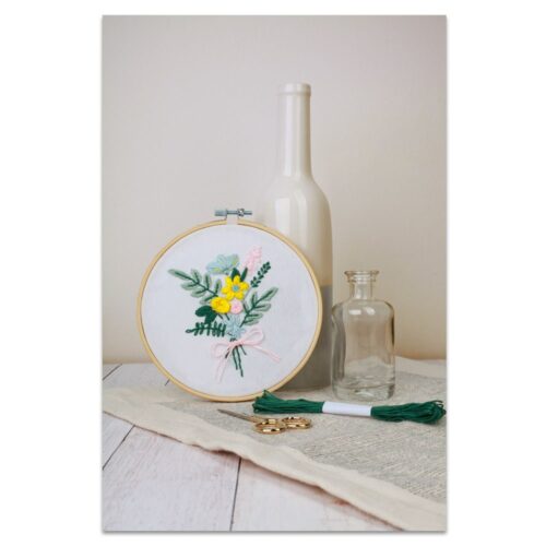 Stickrahmen mit Blumenmuster, neben zwei Glasflaschen und Stickgarn auf einem mit Stoff bedeckten Tisch.