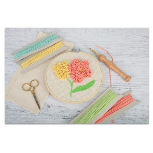Stickrahmen mit buntem Blumenmuster, umgeben von Fäden, Schere und Punch-Needle-Werkzeug.