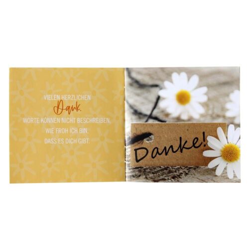 Offene Karte mit Gänseblümchen und einem Schild mit der Aufschrift „Danke!“ auf Holzuntergrund. Deutscher Text auf der linken Seite.