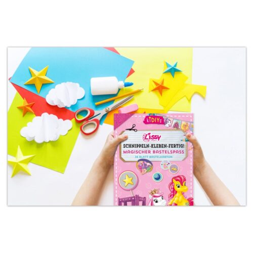 Ein buntes Bastel-Set mit Papier, Schere, Kleber und einem rosa Bastelbuch mit Sternen und Einhörnern.