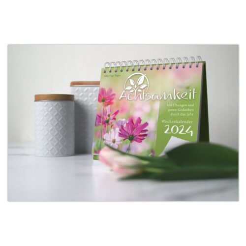 Ein Achtsamkeitskalender für 2024 auf einem Tisch mit rosa Blumen und dekorativen Gläsern.