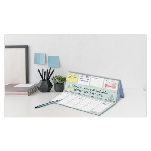 Schreibtisch mit Kalender, Stift, geometrischen Blumentöpfen und einem Stapel Papiere vor einer weißen Wand.