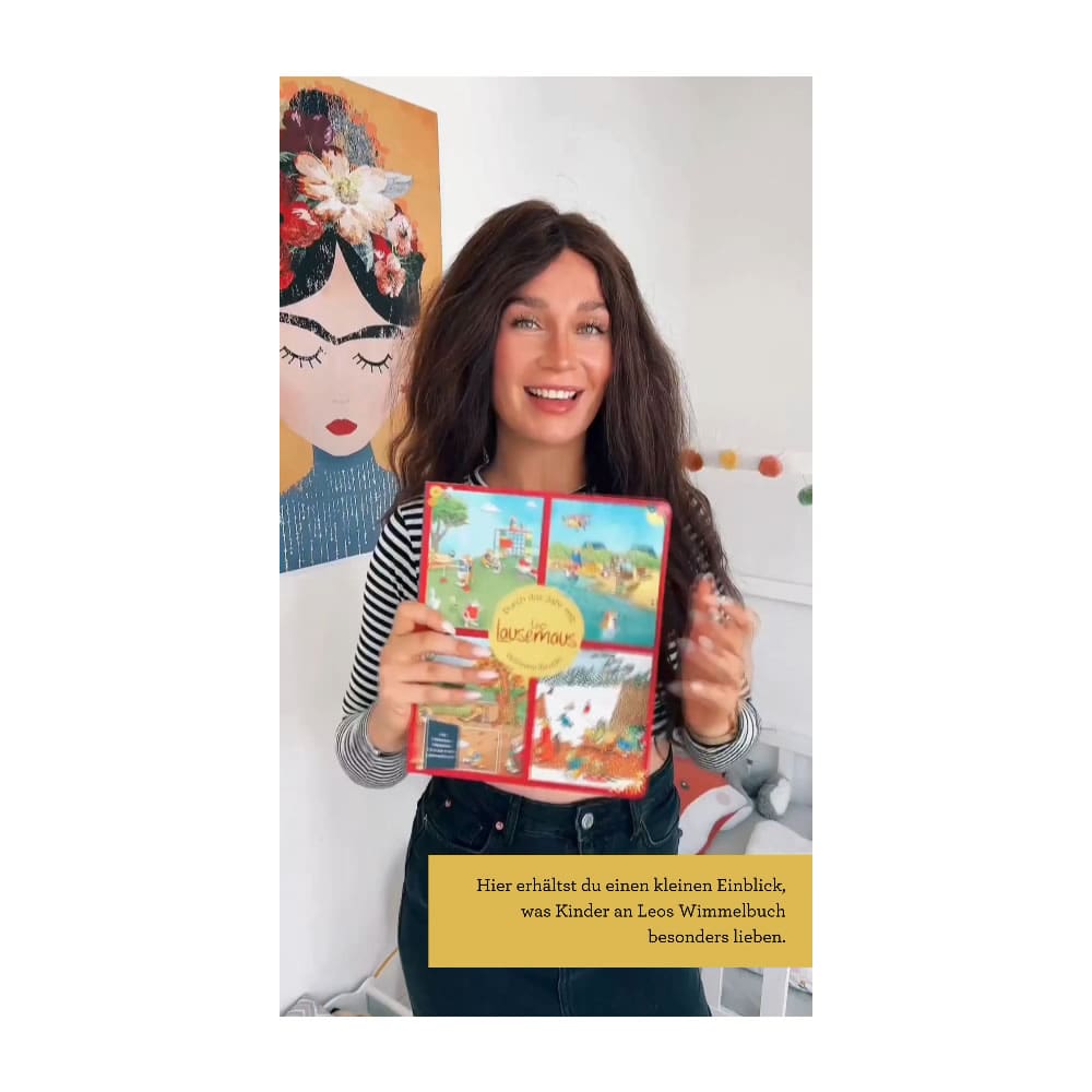 Lächelnde Frau hält ein buntes Kinderbuch vor einer Wand mit einem illustrierten Gesicht.